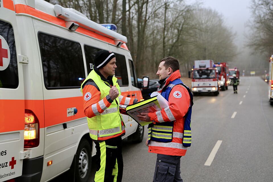 Das Bild zeigt zwei Einsatzkräfte des Roten Kreuzes, die vor einem Einsatzfahrzeug stehen. Im Hintergrund sind weitere Einsatzfahrzeuge zu sehen. 