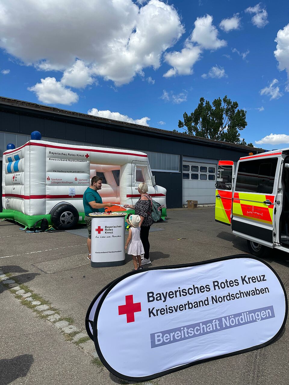 Das Bild zeigt im Hintergrund die Rettungswagen-Hüpfburg des BRK-Nordschwaben. Vor der Hüpfburg steht ein Infostand, an dem ein JRK-Mitglied einer interessierten Passantin Informationen zum Roten Kreuz gibt. Im Vordergrund sieht man das Wurfbanner der BRK-Bereitschaft Nördlingen. 