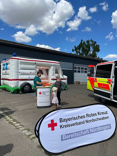 Das Bild zeigt im Hintergrund die Rettungswagen-Hüpfburg des BRK-Nordschwaben. Vor der Hüpfburg steht ein Infostand, an dem ein JRK-Mitglied einer interessierten Passantin Informationen zum Roten Kreuz gibt. Im Vordergrund sieht man das Wurfbanner der BRK-Bereitschaft Nördlingen. 