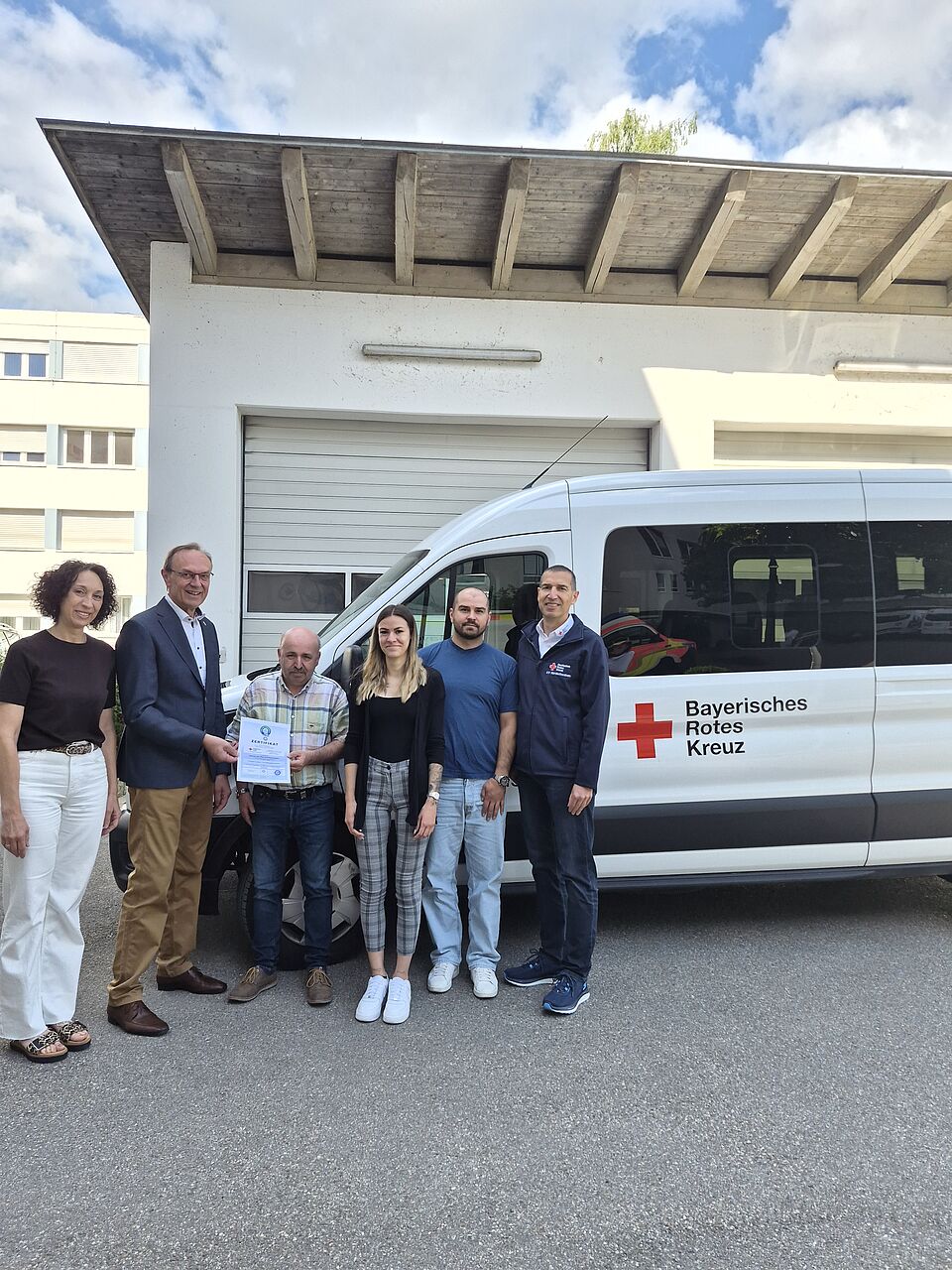 Das Bild zeigt von links nach rechts vor einem Fahrzeug des Fahrdienstes stehend Nicole Uhl (BRK-Fahrdienst), Johann Natzer (BRK-Kreisvorsitzender), Norbert Schuster (Sachgebietsleiter BRK-Fahrdienst), Viviana Popolo (BRK-Fahrdienst), Viktor Nau (BRK-Fahrdienst), Arthur Lettenbauer (BRK-Kreisgeschäftsführer). Herr Natzer übergibt das Zertifikat an Herrn Schuster.