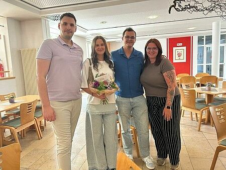 Auf dem Bild sind v.l.n.r. folgende Personen zu sehen: Ismet Krekic (stv. PDL), Nikolina Vidovic, Mihael Vidovic, Celina Sauerland (Praxisanleiterin). Sie stehen im Foyer des BRK-Pflegezentrums am Mangoldfelsen in Donauwörth. Nikolina Vidovic hat einen Blumenstrauß in der Hand. 