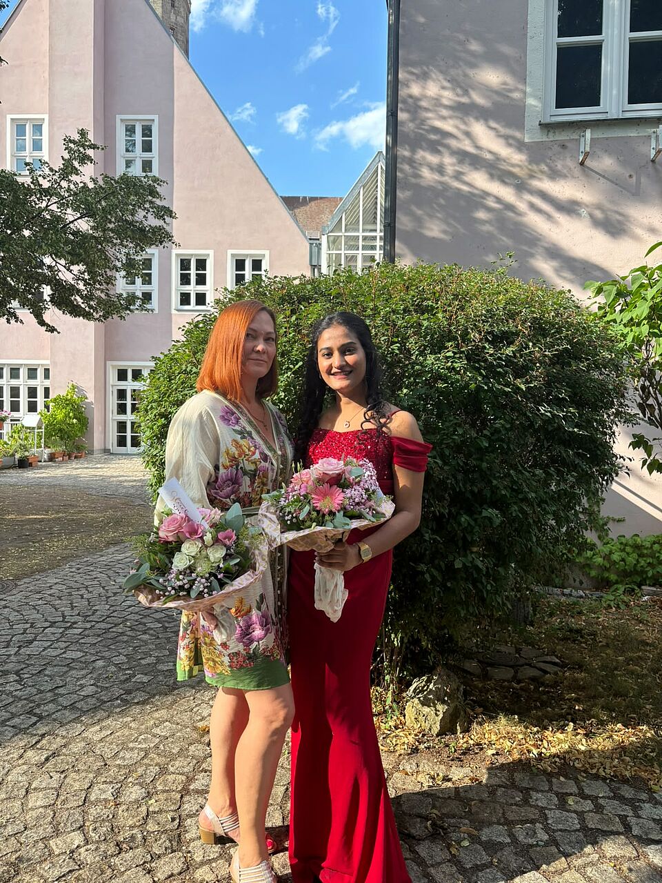 Auf dem Bild sind von links nach rechts Hanna Rott und Rinta Kochuparambil Manoj zu sehen. Beide stehen vor einem grünen Buchsbusch und haben jeweils einen Blumenstrauß in der Hand. 