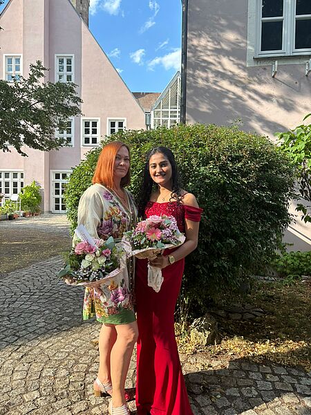 Auf dem Bild sind von links nach rechts Hanna Rott und Rinta Kochuparambil Manoj zu sehen. Beide stehen vor einem grünen Buchsbusch und haben jeweils einen Blumenstrauß in der Hand. 