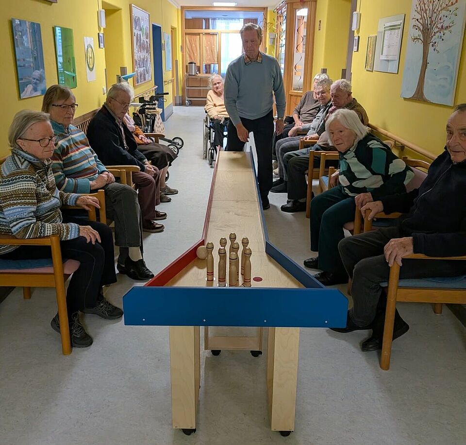 Das Bild zeigt Tagespflegegäste, die die neue mobile Kegelbahn testen. Sie sitzen zu beiden Seiten an der Kegelbahn. Ein Gast steht vor ihr und hat die Kugel geworfen, um die Kegel umzustoßen. 